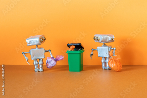 Two small silver metal robots are holding garbage bags ready to be disposed of in a dumpster, trash can. Orange background.