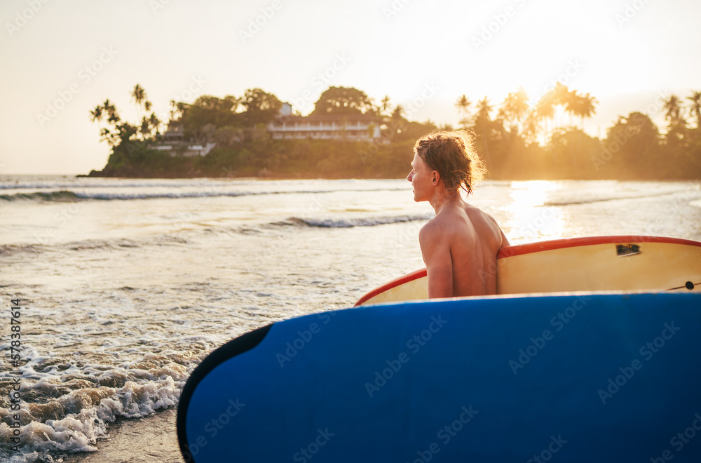 Young teenage boy with old surfboard goes to the sea for surfing. He enjoying a beautiful sunset light on Dewata beach on Sri Lanka. Active sport vacation and exotic countries traveling concept.