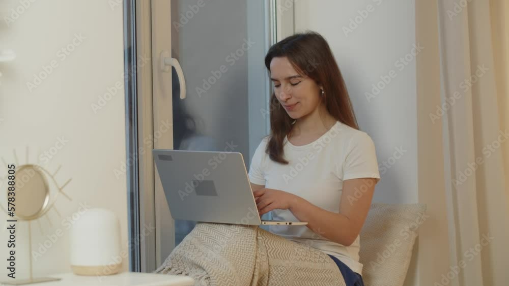Smiling woman chatting with friends using a laptop. A girl is sitting on the windowsill in her room and using computer. Working from home. Smiling woman chatting with friends using a laptop. A girl is sitting on the windowsill in her room and using computer. Working from home.