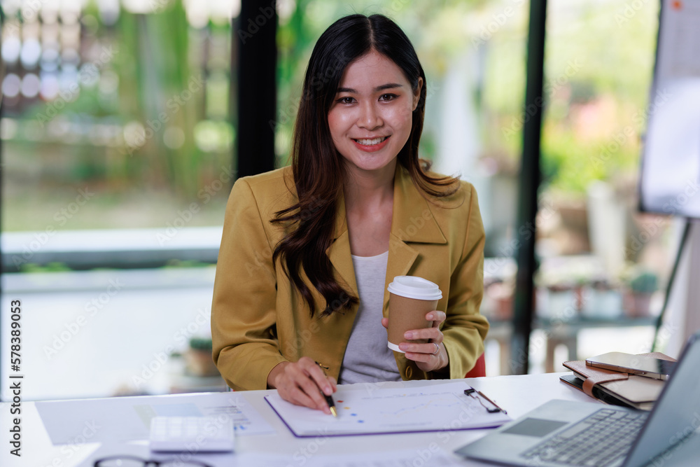 Concept of business office woman working,Businesswoman smile while working about her invesment plan with analyzing document and business investment graph data and holding coffee cup in workstation Concept of business office woman working,Businesswoman smile while working about her invesment plan with analyzing document and business investment graph data and holding coffee cup in workstation