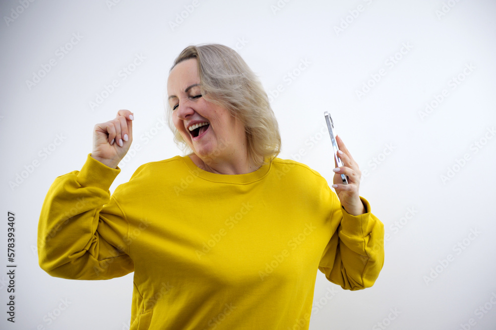 middle-aged adult woman dancing in bright clothes in studio in the hands of a phone with a white screen space for text advertising positive emotions middle-aged adult woman dancing in bright clothes in studio in the hands of a phone with a white screen space for text advertising positive emotions