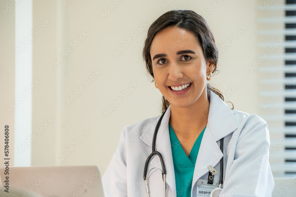 Woman female doctor portrait in white coat gown with stethoscope sit and smile to front. Medical student attractive friendly wear white coat smiling and work on table in patient room Woman female doctor portrait in white coat gown with stethoscope sit and smile to front. Medical student attractive friendly wear white coat smiling and work on table in patient room