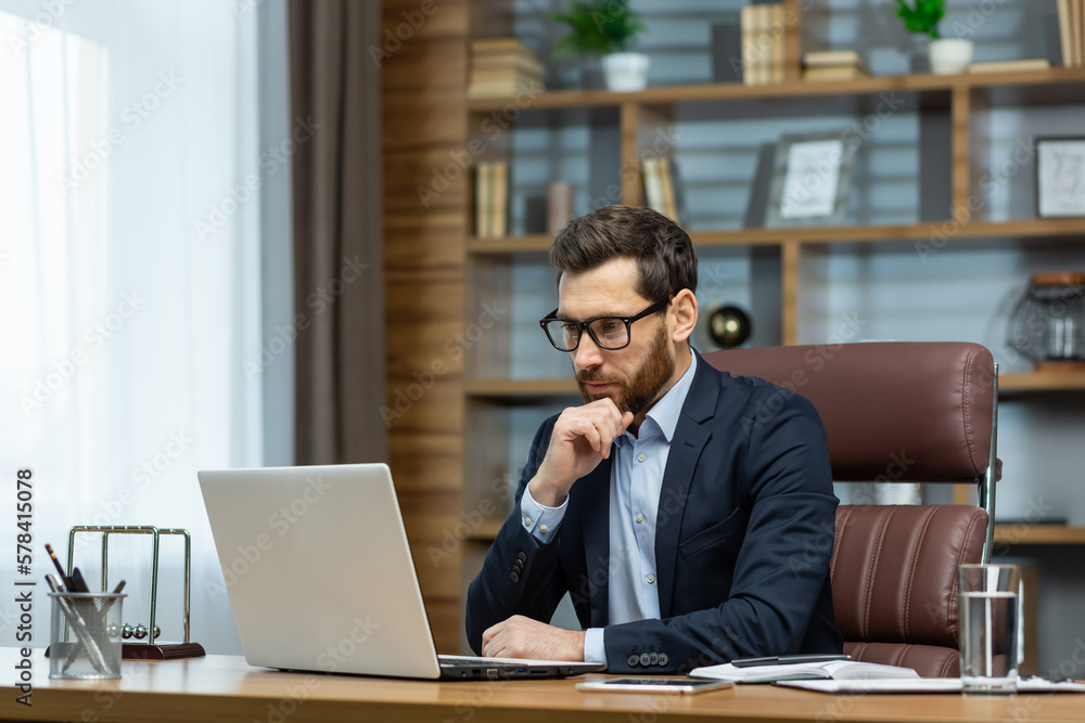 Thinking businessman working sitting at desk, mature adult boss in business suit and beard looking at laptop screen thinking about financial investment decisions inside office. - obrazy, fototapety, plakaty 