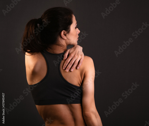 Female sporty muscular with ponytail doing stretching workout of the shoulders, blades in sport bra, holding the neck the hand on dark background with empty copy space. Back view.