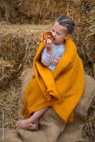 A girl wrapped in a yellow cloth eats a cinnamon bun and nuts against the background of straw bales