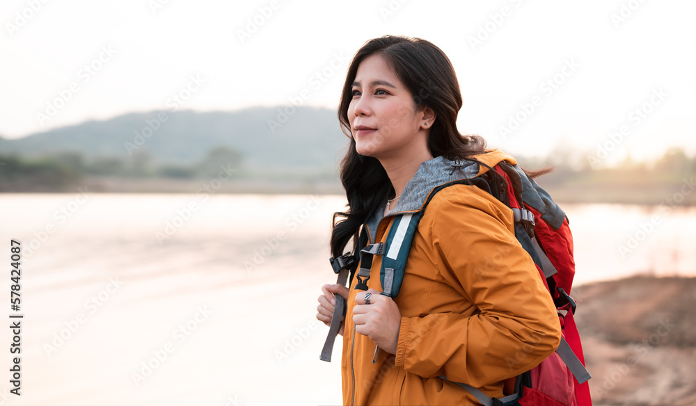 Beautiful asian woman backpacker with backpack travelling for camping near the lake, smiling female traveller wearing yellow jacket relaxing with nature on vacation. Beautiful asian woman backpacker with backpack travelling for camping near the lake, smiling female traveller wearing yellow jacket relaxing with nature on vacation.