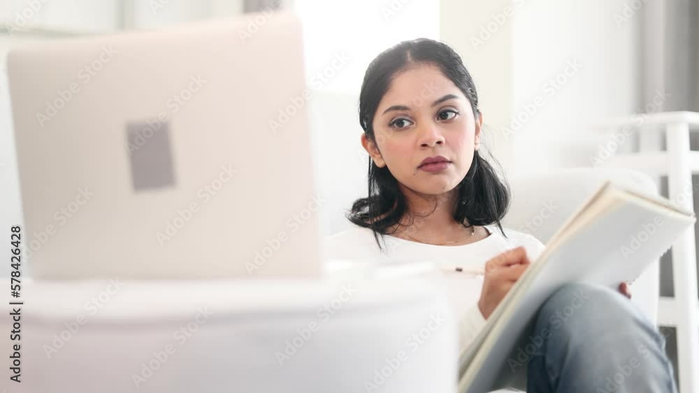 Portrait of focused indian girl preparing for exam and studying at home Charming student concentrated on learning and make notes in her notebook while looking at laptop computer screen indoors Portrait of focused indian girl preparing for exam and studying at home Charming student concentrated on learning and make notes in her notebook while looking at laptop computer screen indoors
