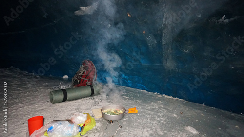 A guy cooks food on gas inside an ice cave. The climber lights the burner, cuts sausage and tomato. Breaks the eggs and fry in a frying pan. Steam from food. The ice wall is blue and the floor is gray