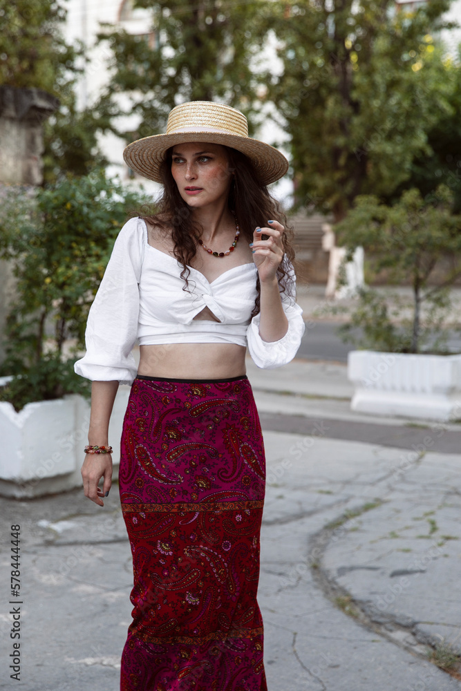 The beautiful brunette with curly hair, wearing a straw hat and a white blouse and skirt, poses outside.
