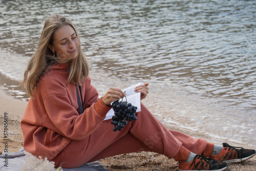 A girl in an orange tracksuit, hoodie and pants sits on the beach and enjoys. Sits by the sea and eats fresh bread, fruits, grapes and reads a book. High quality photo photo