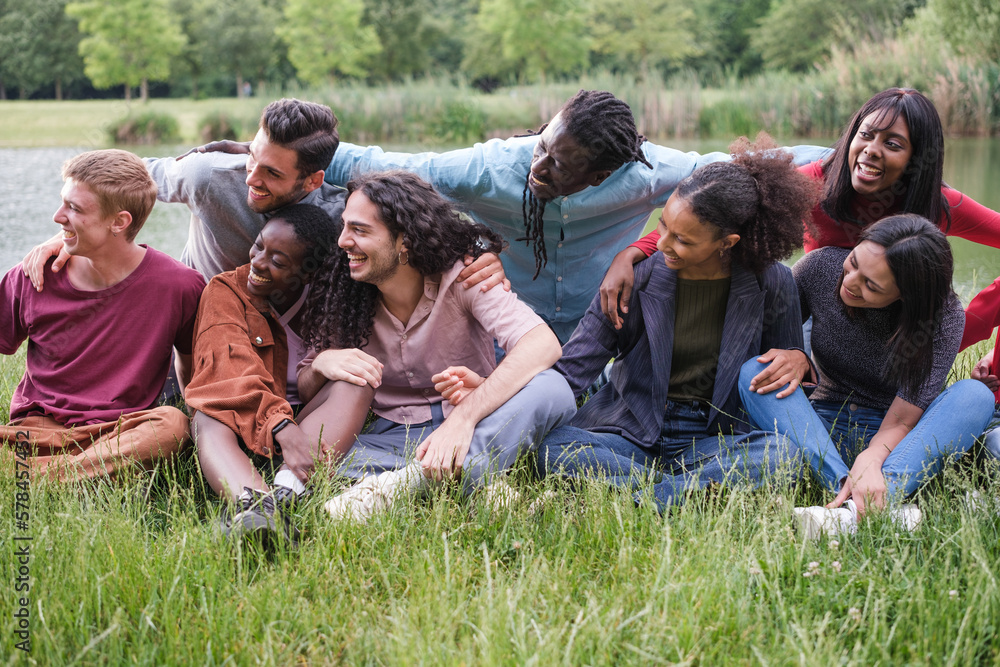 Group of multicultural young friends sitting on the grass by the river in a sunset outdoors. Concept: Lifestyle, diversity, friendship Group of multicultural young friends sitting on the grass by the river in a sunset outdoors. Concept: Lifestyle, diversity, friendship