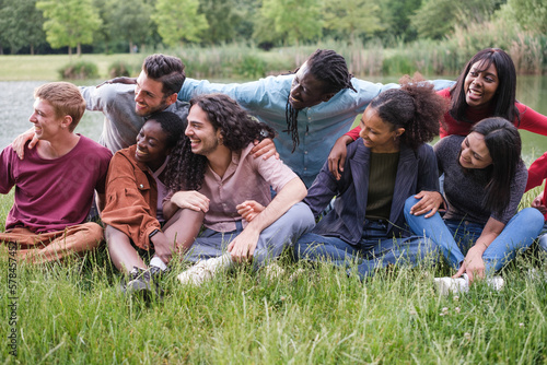 Group of multicultural young friends sitting on the grass by the river in a sunset outdoors. Concept: Lifestyle, diversity, friendship