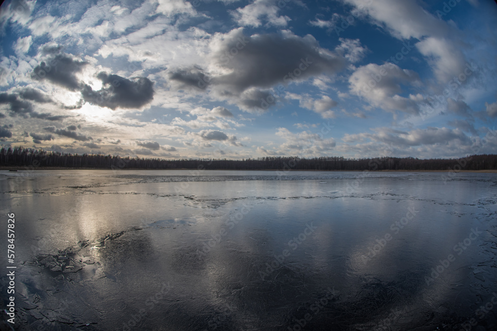 Evening landscape of a spring lake covered with thin cracking transparent ice and a cloudy sky reflected in it. Fish eye.