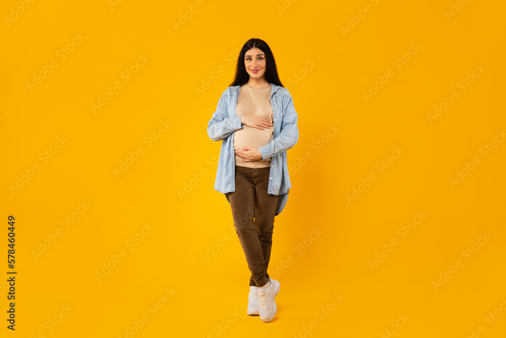 Full length shot of happy pregnant woman embracing belly and smiling at camera, posing on yellow studio background Full length shot of happy pregnant woman embracing belly and smiling at camera, posing on yellow studio background