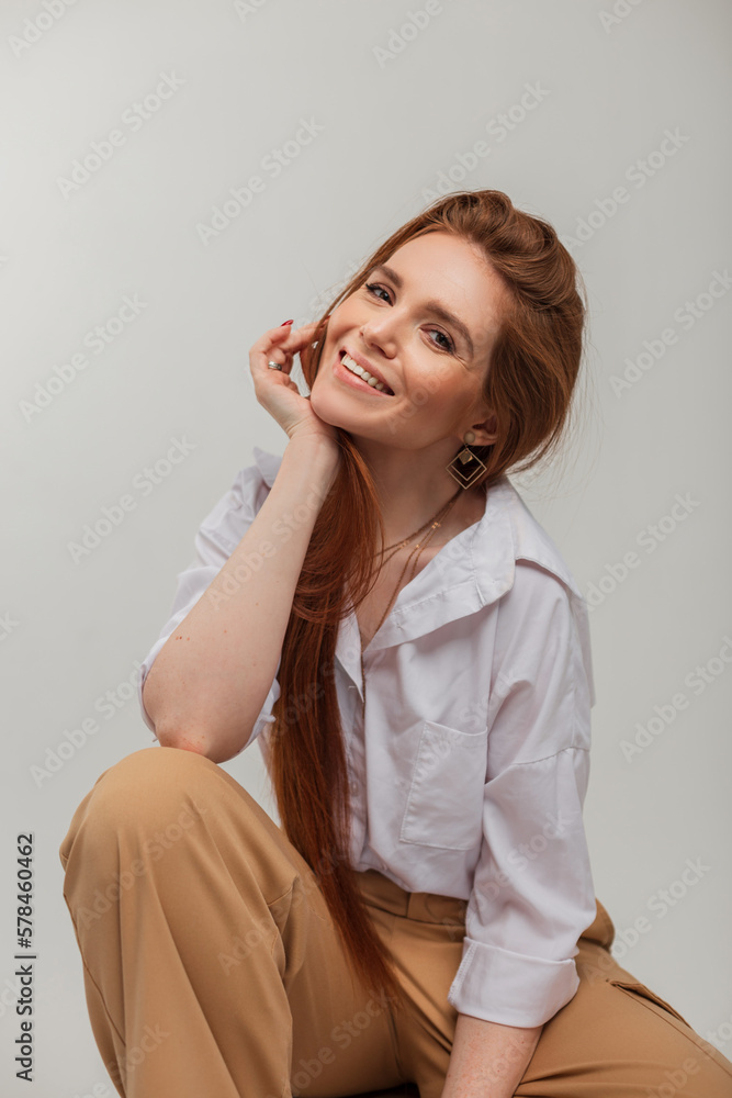 Natural female portrait of a beautiful cheerful cute woman with a beautiful smile in fashion clothes with trousers and a shirt sits on a white background in the studio Natural female portrait of a beautiful cheerful cute woman with a beautiful smile in fashion clothes with trousers and a shirt sits on a white background in the studio