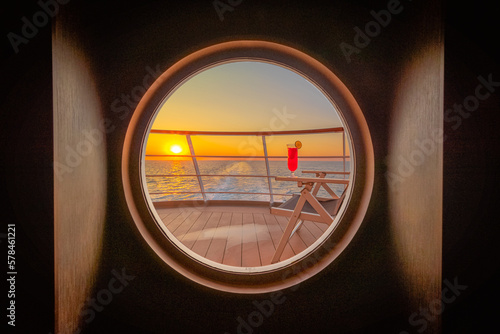 Pont promenade d'un navire de croisière en navigation avec un transat et un cocktail rouge au coucher de soleil à travers un hublot. 