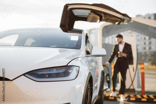 Blurred background of indian man in formal wear restoring battery of his electric car at EV charging station. Focus on white shiny luxury auto. Eco friendly transport. © sofiko14
