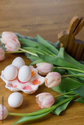 Happy Easter! Beautiful tulips, eggs and bunny decoration on wooden table. Modern farmhouse easter decor. Stylish handmade egg holder, natural eggs, pink tulips and rustic bunny