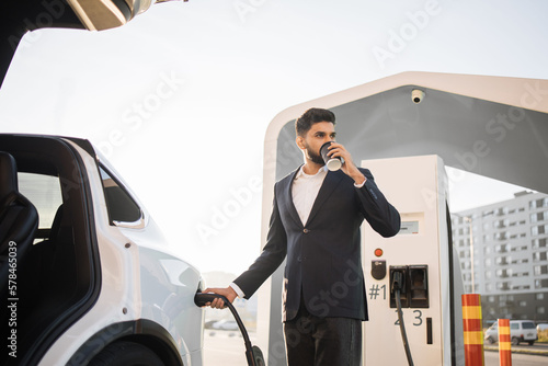 Well-dressed arabian man enjoying hot aroma coffee while charging battery of white electric car at urban station. Concept of consciousness and eco friendly auto.