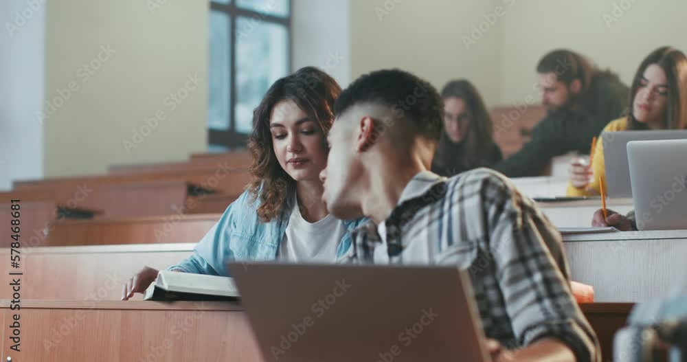 Caucasian beautiful girl reading textbook at lection and asking something guy with laptop. Whispering at seminar in college. University study concept. Female read book at class and talking. Students. Caucasian beautiful girl reading textbook at lection and asking something guy with laptop. Whispering at seminar in college. University study concept. Female read book at class and talking. Students.