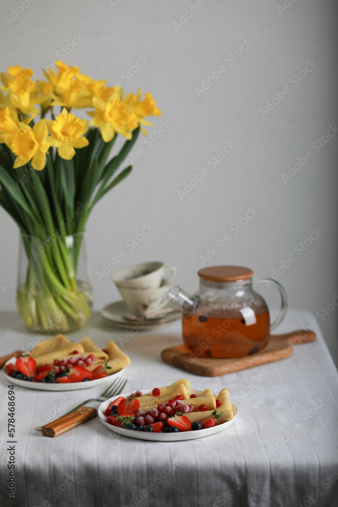 Thin pancakes with strawberries and blueberries, delicious breakfast. Homemade french crepes with fresh fruits, teapot with berries tea and bouquet of yellow flowers on table. Thin pancakes with strawberries and blueberries, delicious breakfast. Homemade french crepes with fresh fruits, teapot with berries tea and bouquet of yellow flowers on table.