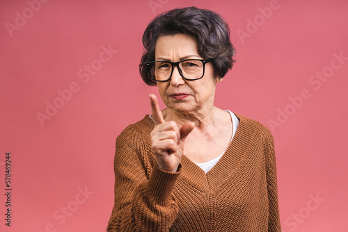 Portrait of angry grey haired old strict senior woman wearing glasses pointing up threatening with finger. Grandmother isolated over pink background.