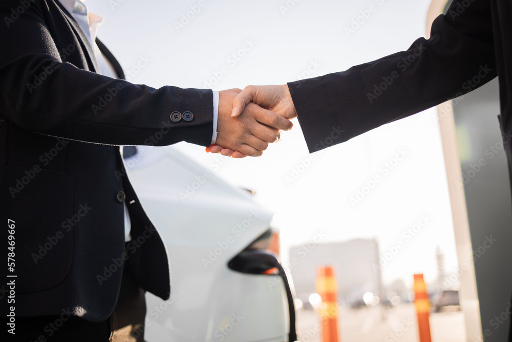 Fototapeta premium Close up of caucasian woman and asian dealership seller shaking hands in front of white luxury electric car. Concept of people, good deal and new transport.