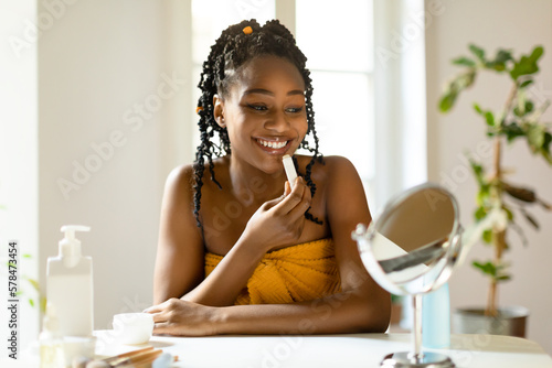 Foto Happy beautiful black woman using moisturizing lipbalm, lady sitting at vanity t