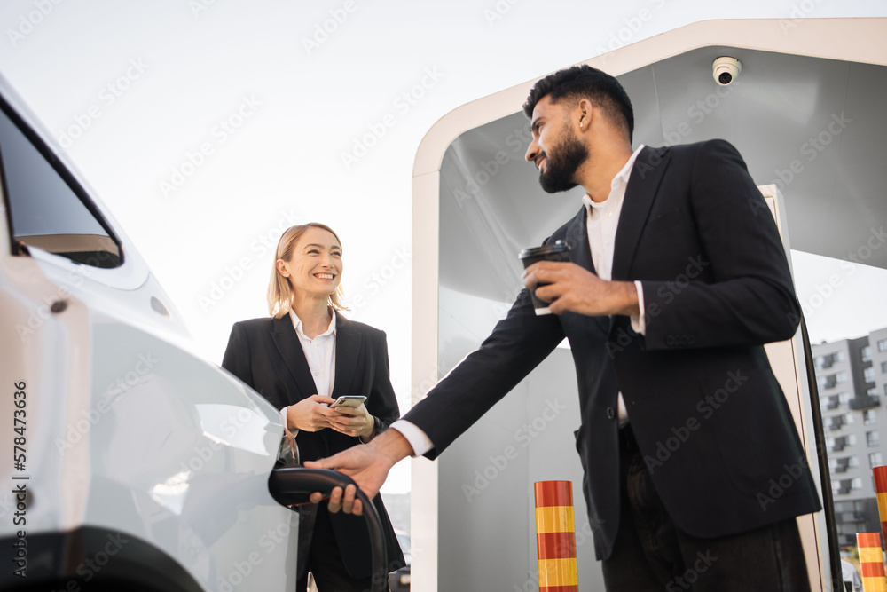 Smiling caucasian woman and handsome indian man dressed in formal wear using smartphone and drinking coffee on EV station. Two business colleagues chatting about work while charging car. Smiling caucasian woman and handsome indian man dressed in formal wear using smartphone and drinking coffee on EV station. Two business colleagues chatting about work while charging car.