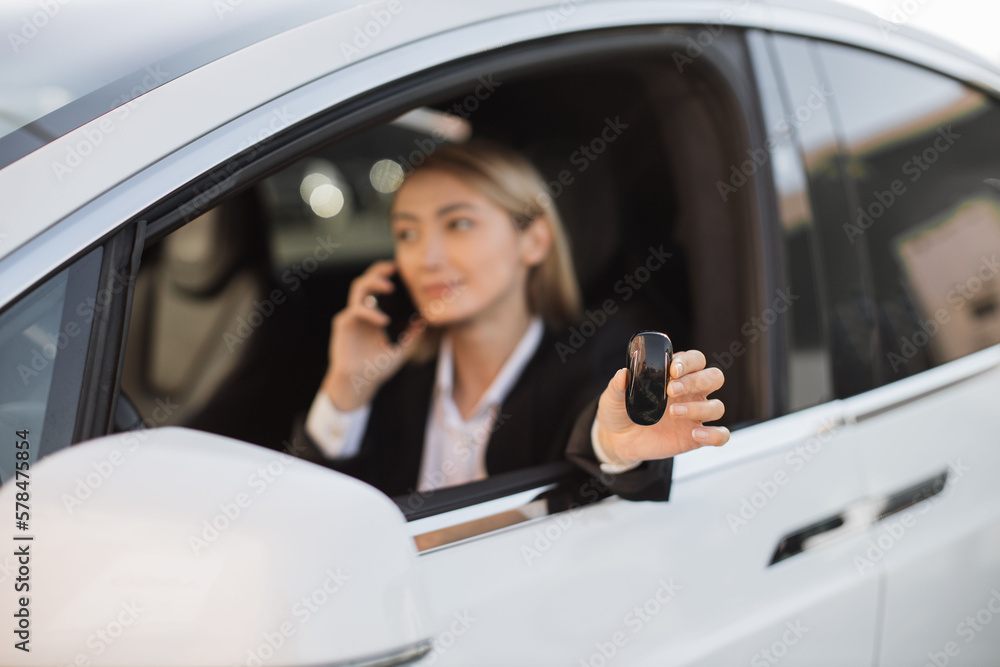 Confident caucasian woman in formal attire talking on mobile phone and holding keys from her luxury new auto. Beautiful young female enjoying expensive purchase at modern shop.