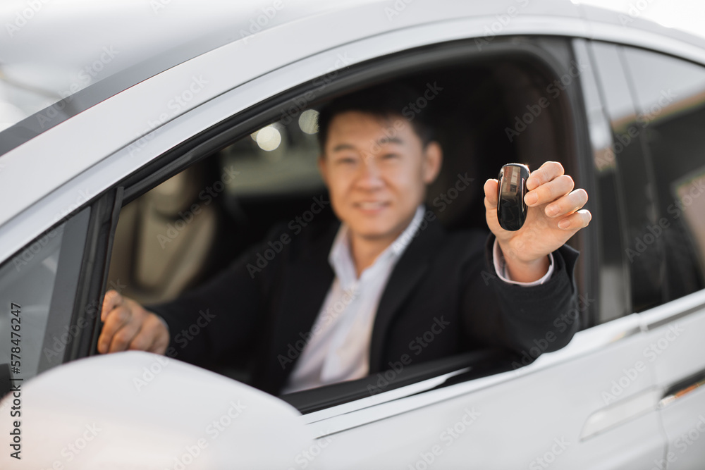 Blurred background of smiling asian businessman in black suit sitting inside new auto with open window. Focus on male hand holding car keys. Transport purchasing concept.