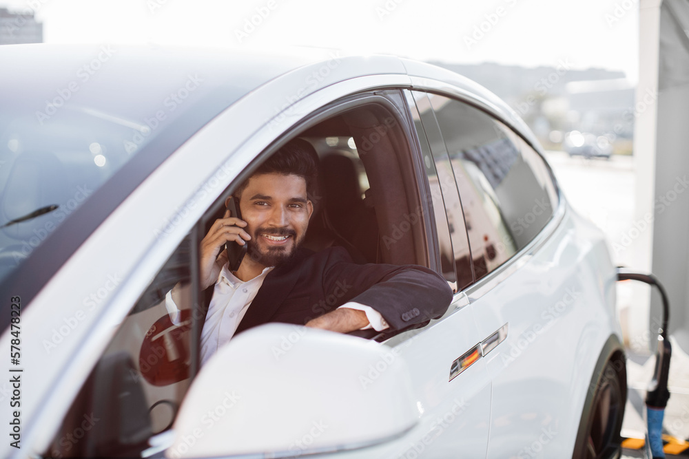 Attractive bearded man in formal suit talking on smartphone while sitting inside brand new auto. Well-dressed indian man smiling happily while looking out of open car window outdoors. Attractive bearded man in formal suit talking on smartphone while sitting inside brand new auto. Well-dressed indian man smiling happily while looking out of open car window outdoors.