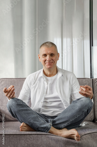 Portrait of a man who sits on a sofa in a room in a lotus position. © Dzmitry