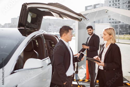 Focused multiethnic coworkers in formal clothing holding modern gadgets in hands. Arabic colleague with takeaway coffee supplying battery-drive vehicle with energy at public charging point.