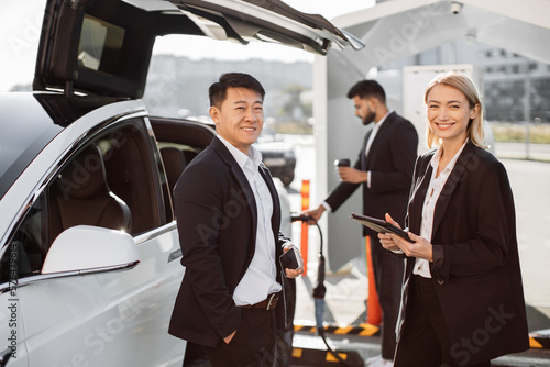 Relaxed smiling multiracial business colleagues standing at e-car charging point with tablet and mobile in hands. Indian man holding takeaway cup and getting auto filled up with electricity behind.