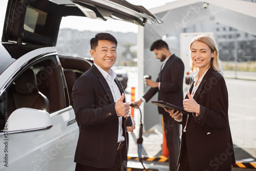 Relaxed smiling multiracial business colleagues standing at e-car charging point with tablet and mobile in hands showing thumbs up.