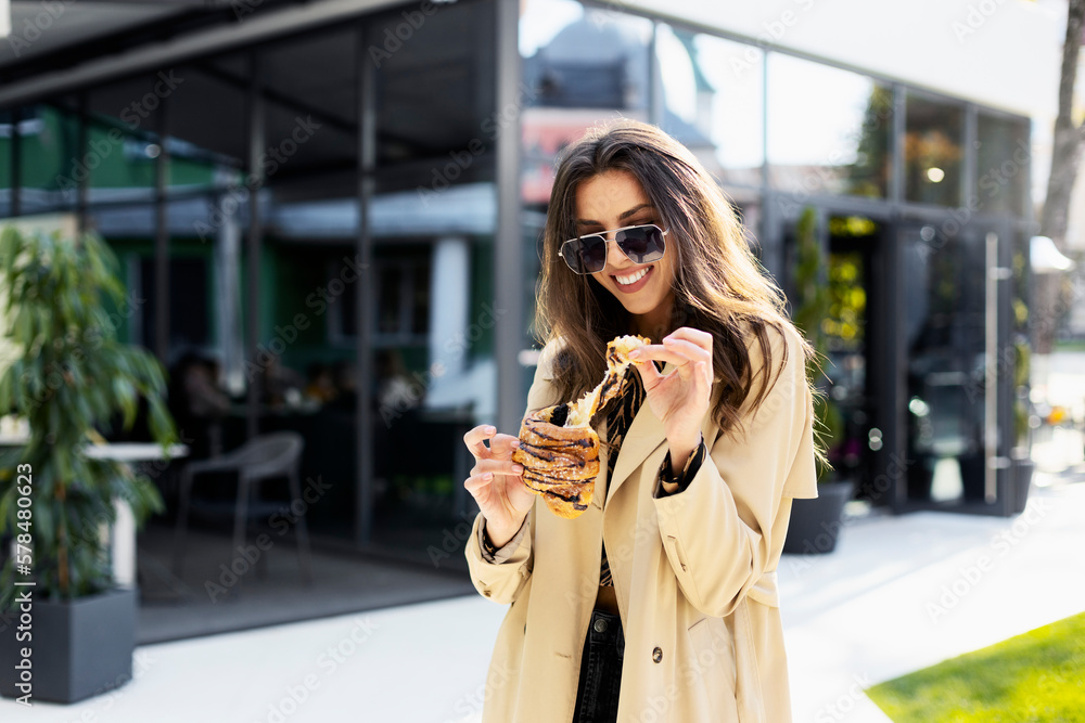 Smiling girl breaks off a piece of croissant. An attractive girl in sunglasses eats a croissant against the background of a modern building, on the street.
