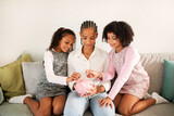Black Mother And Daughters Putting Coins In Piggybank At Home