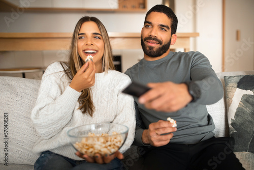 Glad millennial arab guy with beard and european female watch TV  eat popcorn  enjoy movie together on sofa