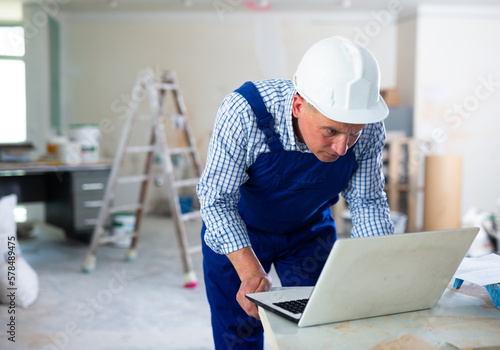Foreman in overall using laptop in construction site, repair works in apartment.