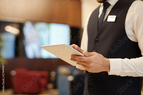 Close-up of young elegant male receptionist using tablet while standing in front of camera in lounge of modern luxurious hotel during work