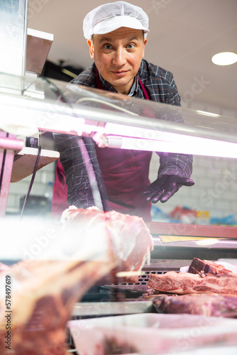 Positive man, professional butcher shop seller, arranging meat products in display case, showing piece of fresh raw veal tenderloin