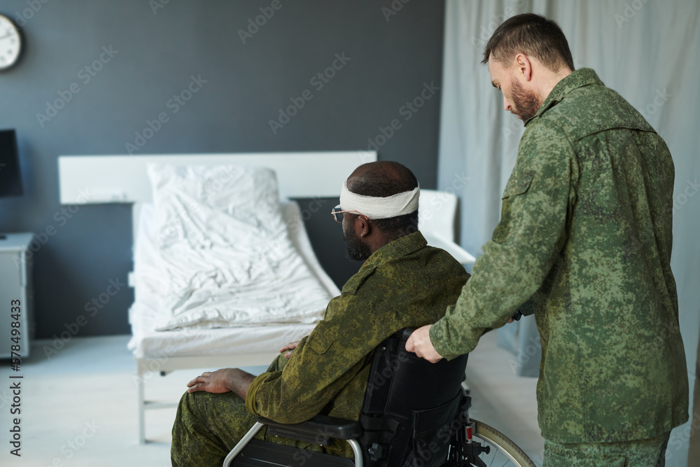 Young soldier in camouflage pushing wheelchair with his friend while taking care of him during course of medical therapy after injury