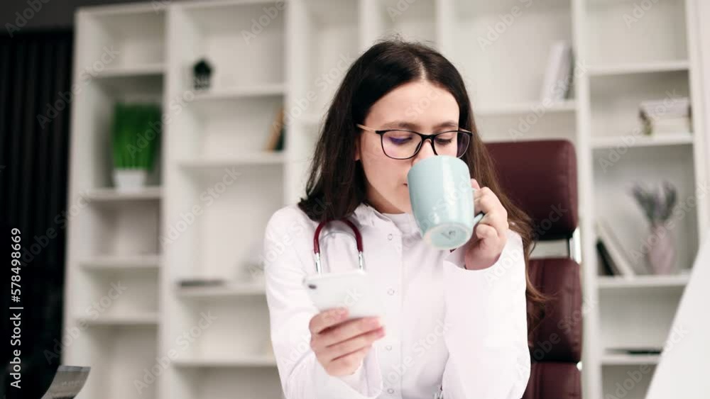 Smiling young doctor therapist with stethoscope have break relax with cup of coffee or tea hold smartphone scrolling social feed has chat with patient or using medical app at light workplace Smiling young doctor therapist with stethoscope have break relax with cup of coffee or tea hold smartphone scrolling social feed has chat with patient or using medical app at light workplace