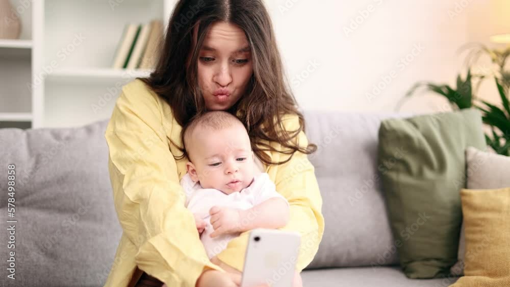 Portrait of cute young woman mother hug her little baby and grimacing taking selfie photograph saving great memories on smartphone mobile phone at light home Happy motherhood concept Portrait of cute young woman mother hug her little baby and grimacing taking selfie photograph saving great memories on smartphone mobile phone at light home Happy motherhood concept