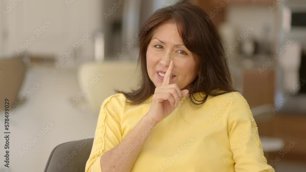 Stylish attractive woman yellow top looking at camera laughing smiling making humorous hand gesture to shush be quiet putting finger to lips sitting in kitchen chair. Mature female asian is happy. Stylish attractive woman yellow top looking at camera laughing smiling making humorous hand gesture to shush be quiet putting finger to lips sitting in kitchen chair. Mature female asian is happy.
