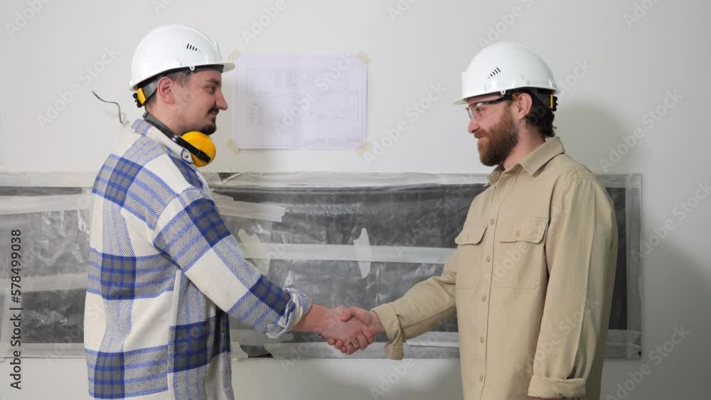 Two young male engineers in white helmets shake hands with a smile and stand in the middle of the room where repair work is underway. High quality 4k footage Two young male engineers in white helmets shake hands with a smile and stand in the middle of the room where repair work is underway. High quality 4k footage