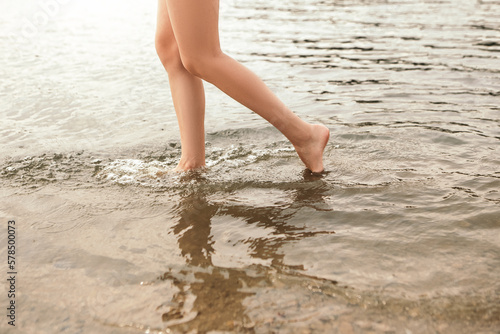 Close up naked slim woman legs walking barefoot along the river. Female feet on sea beach with splashing waves and foam. Girl tourist on vacation. Lady go on shore after bathing