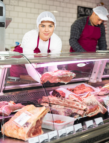 Successful young female butcher shop worker preparing meat products for sale in glass fridge showcase, choosing slice of fresh raw veal sirloin for customer..
