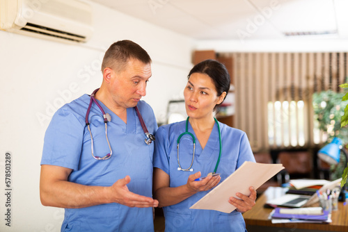 Two professional doctors standing in office, reading medical card of patient and discussing diagnosis